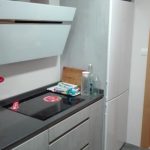 Una combinación de materiales, la zona de arriba blanco brillo y zona de abajo una puerta gris en mate y los decorativos también en gris,con tirador uñero en inox. y encimera de Silestone en color Marengo, vemos la vitrina en cristal blanco con decorado en el mismo color que la encimera.
